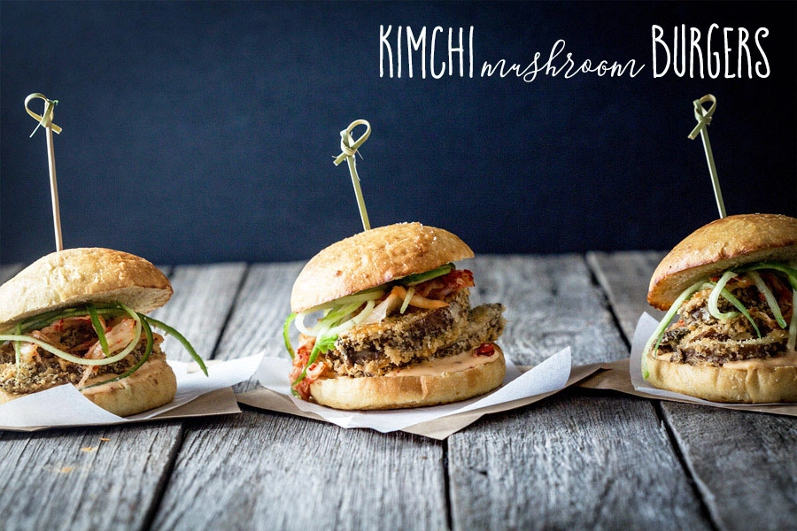 Three vegan kimchi mushroom burgers, side beside, on a rustic wooden table with a dark wall in the background. 