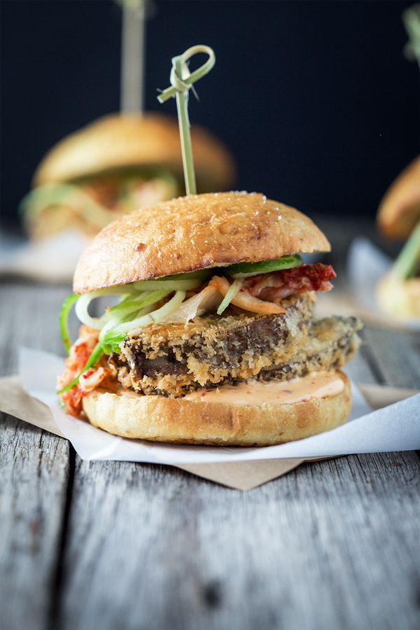 Close up image of a vegan kimchi crispy mushroom burger sitting on white baking paper on a rustic wooden table