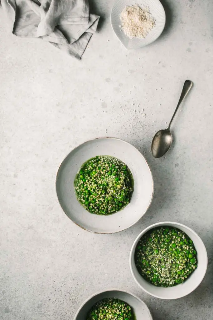 An overhead image of the finished green pea buckwheat risotto served in three white bowls sitting in the bottom right of the image. In the top left corner is a textured napkin and a pinch bowl of pink sea salt.