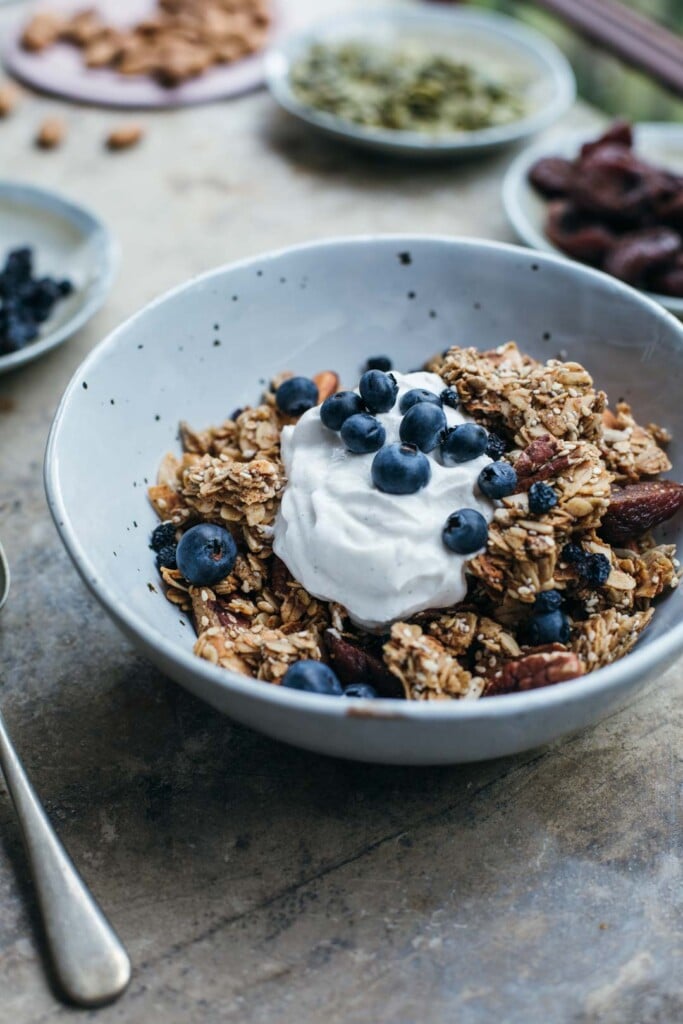 Mid-shot of concrete bench top with a white bowl full of chunky sesame granola in it with coconut yoghurt and blueberries. In the background there are pinch bowls of pepitas, almonds, apricots etc