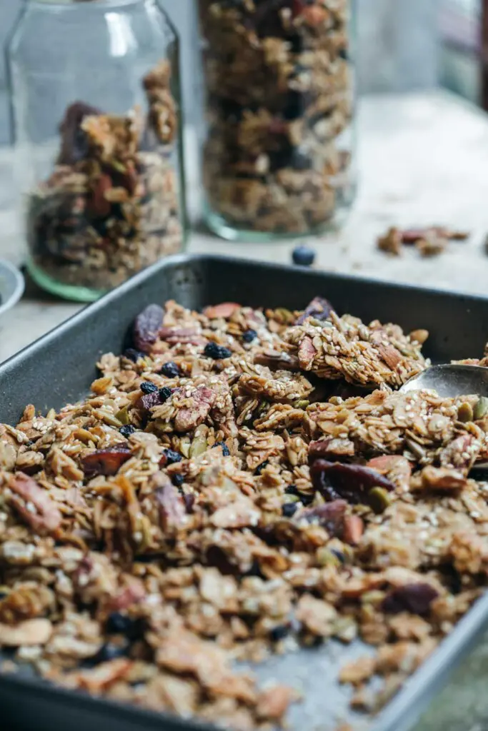 A mid-shot of the tray of freshly baked granola after it has been broken in to chunks. Two jars of granola are in the background. 