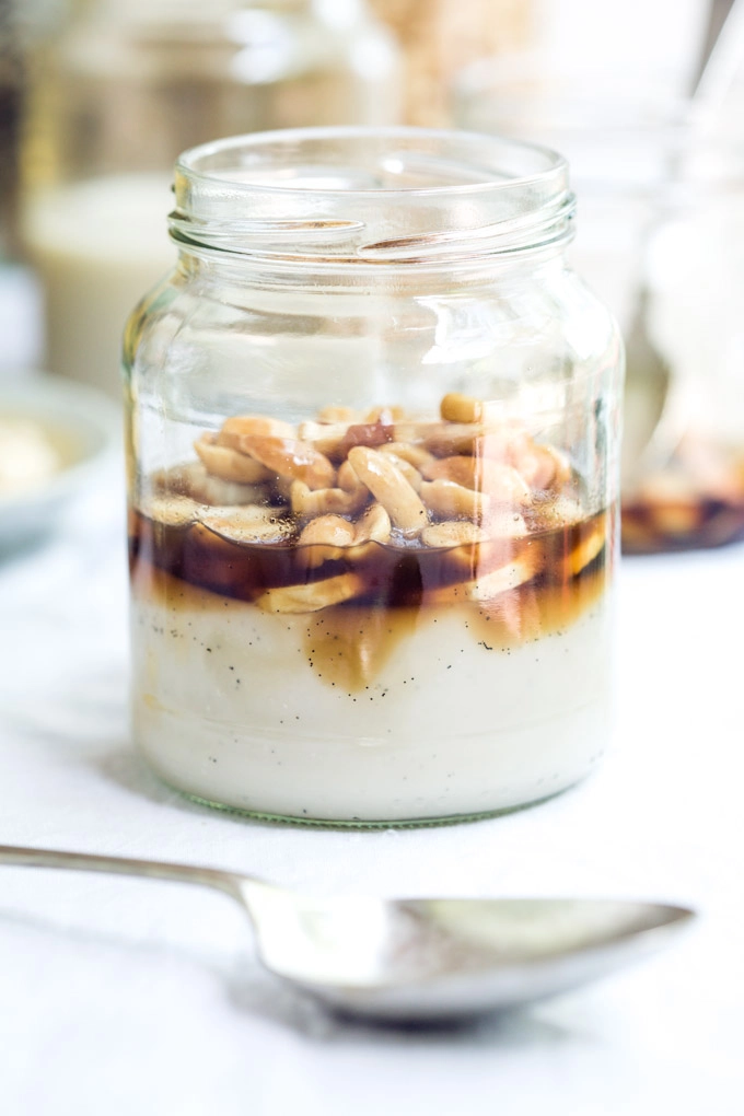 A front-on image of a freshly set vegan panna cotta in a jar sitting on a white table cloth with a spoon in the foreground