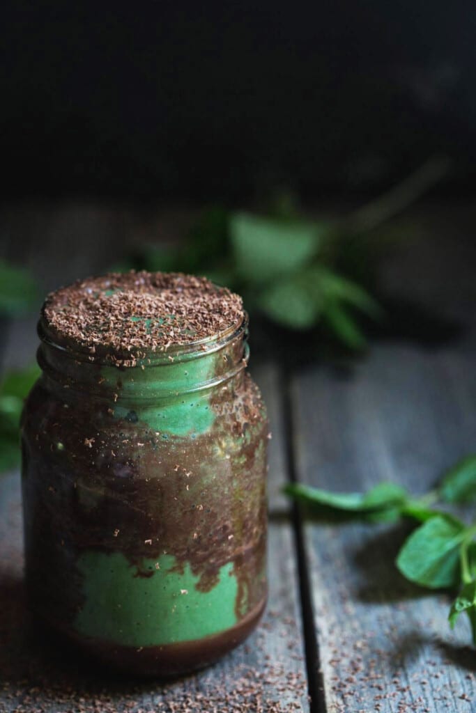 A large jar full of the finished dairy-free chocolate mint smoothie with chocolate ripples and dark chocolate shavings on a rustic wooden bench. 
