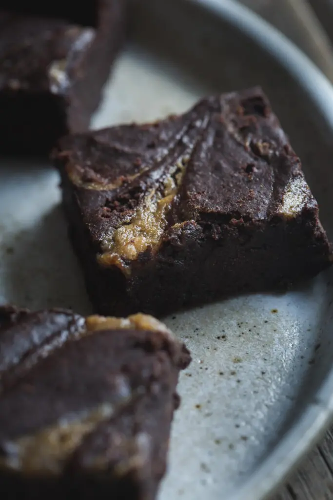 A close-up image of black brownie squares with tahini caramel swirls sitting on a earthenware plate.