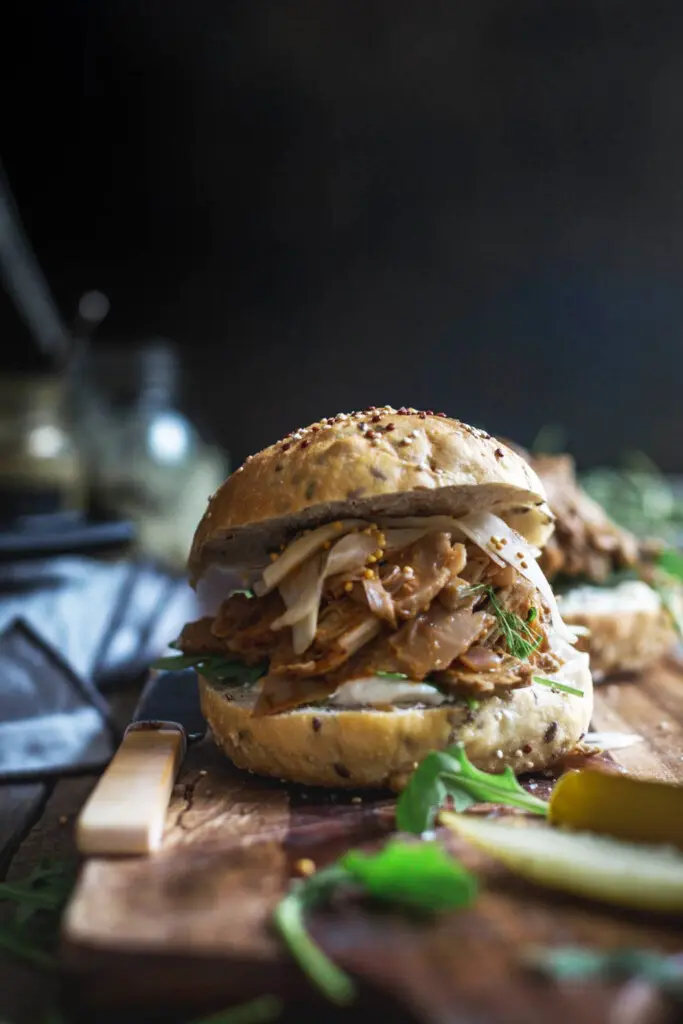 Final image of finished easy Creole jackfruit burgers sitting on a wooden board with jars of pickles in the background 