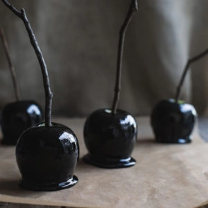 Black toffee apples with twig handles on brown parchment.