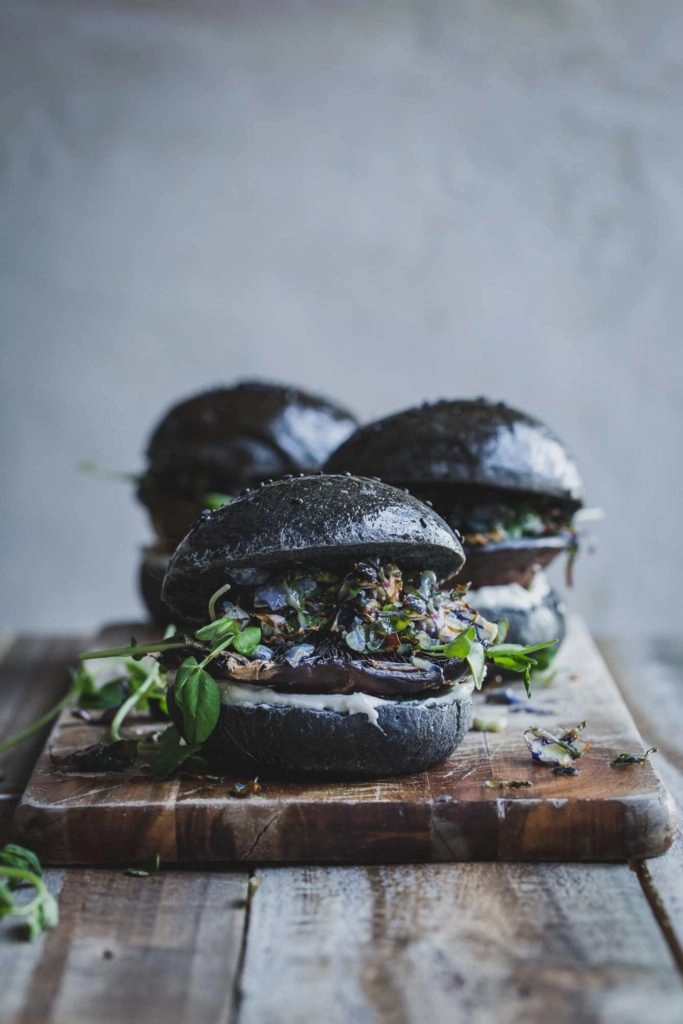 A Portobello Mushroom Burger on a charcoal bun sitting on a wooden board with two burgers in the background slightly to the side