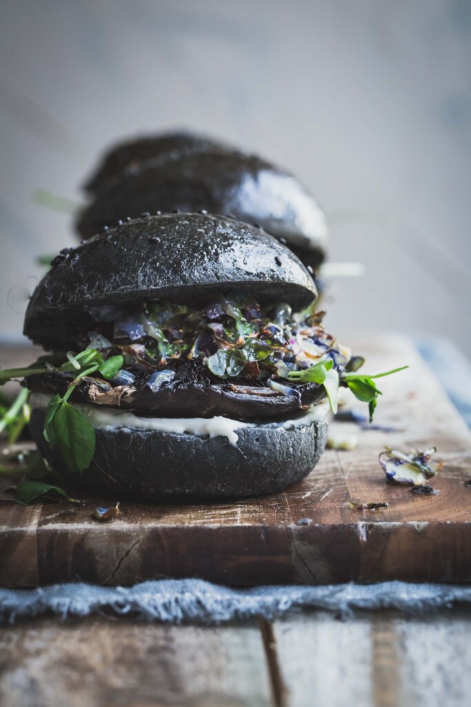 A Portobello Mushroom Burger on a charcoal bun sitting on a wooden board with two burgers in the background