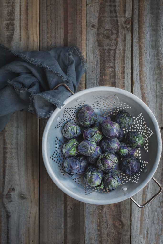Overhead process image of a rustic colander full of freshly washed purple Brussels sprouts sitting on a distressed wooden bench
