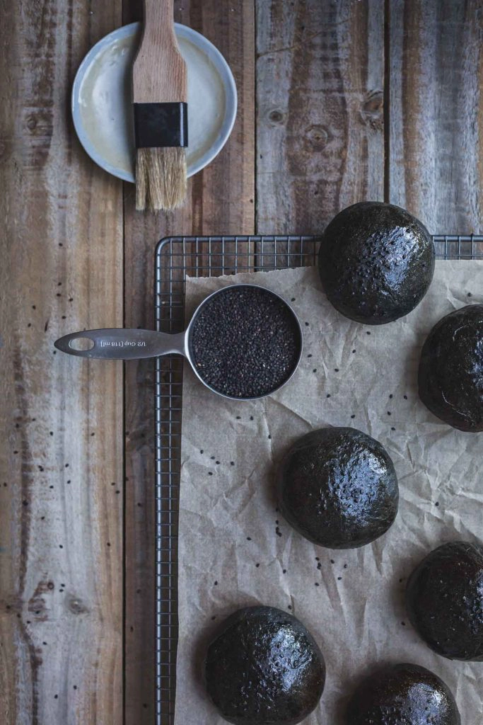 An overhead process shot of a rack of unbaked charcoal buns, a cup of black chia seeds and a pastry brush on a distressed wooden board