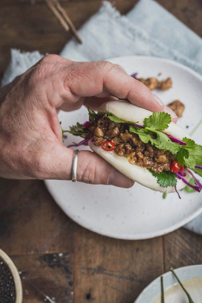 Close-up overhead shot of a man's hand holding a vegan bao bun filled with peking tempeh and greens over a white plate and rustic wooden table. 