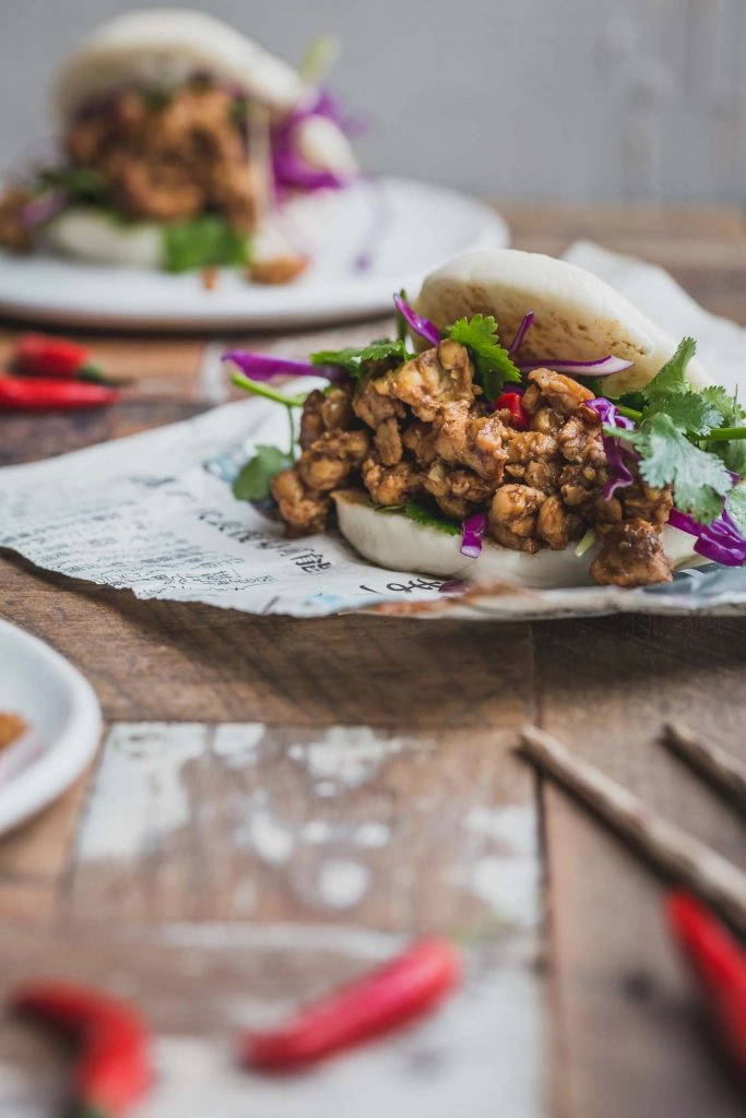 Close up image of a bao bun filled with peking tempeh and greens sitting on a sheet of Asian newspaper. Another bun sits on plate in the background. A pair of chopsticks in the foreground on a rustic wooden table. 