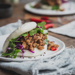 A close up image of a bao bun filled with tempeh and greens on a wooden table.