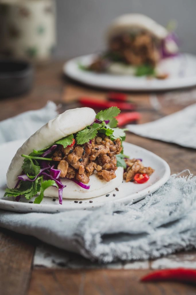 Close up image of a bao bun filled with peking tempeh and greens on a white plate with a grey napkin underneath. Another bun sits in the distance in a rustic wooden table. 