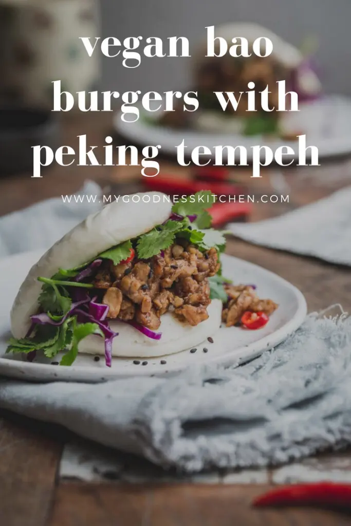 Close up image of a bao bun filled with peking tempeh and greens on a white plate with a grey napkin underneath. Another bun sits in the distance in a rustic wooden table. Title text overlay in white.