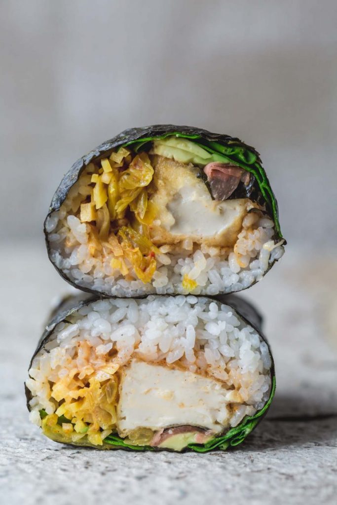 A vegan crispy tofu sushi burrito cut in half and stacked to show inside | sitting on rustic white bench