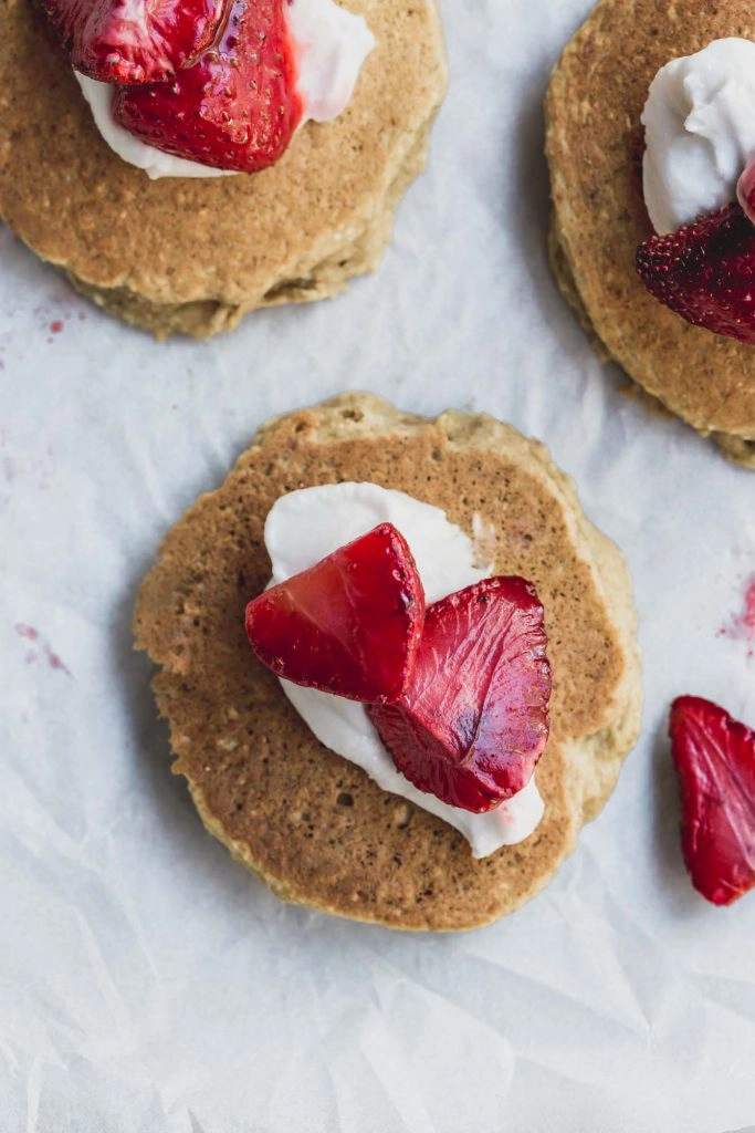 A closeup up image of a banana and coconut pancake topped with cream and roast strawberries.