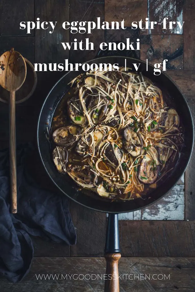 Flat lay image of a large skillet filled with spicy eggplant stir fry with enoki mushrooms next to a wooden spoon on a rustic wooden table with title text overlay