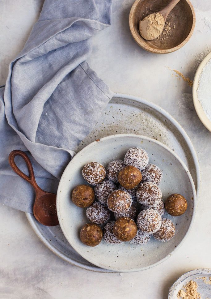 Over-head image of a bowl of almond butter oat energy balls resting on a rustic white plate with a vintage napkin and wooden bowls of ingredients nearby