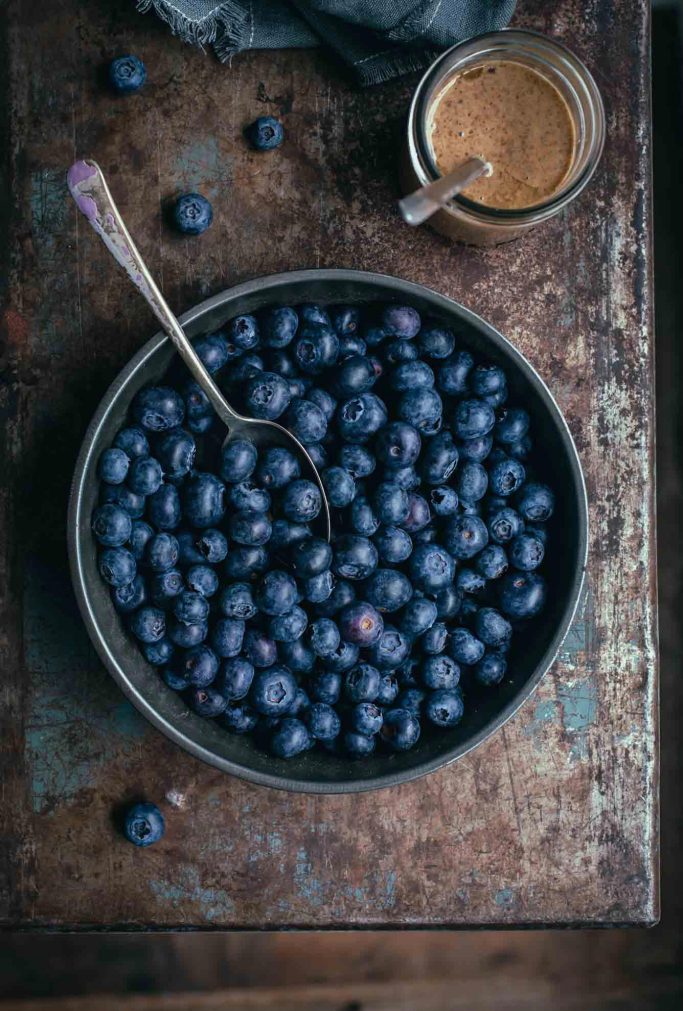 A close-up overhead image of a pie tin filled with fresh blueberries on a rustic rust background with a jar of nut butter off to the side.