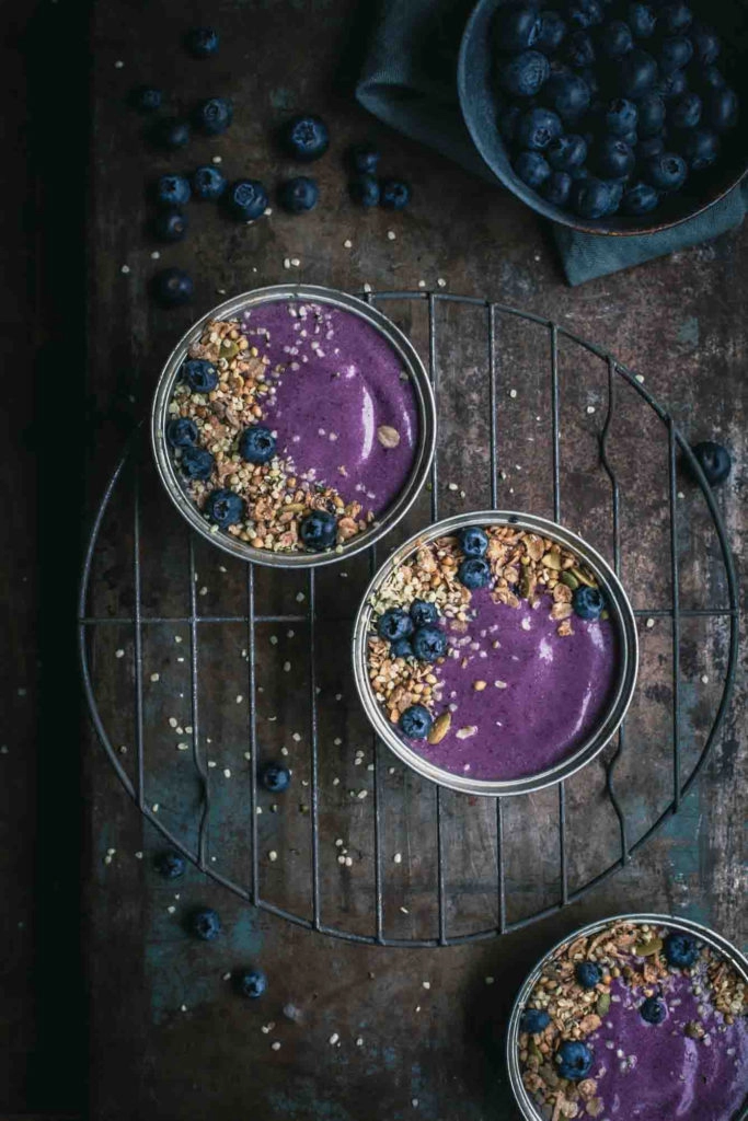 Overhead image of 3 small pie tins filled with thick blueberry pie smoothie, granola and fresh blueberries on a rustic table. A bowl of blueberries sitting off to the side.
