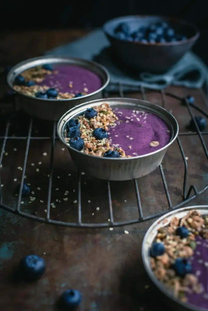 A 45-degree angle image of three small pie tins filled with thick blueberry pie smoothie, granola and fresh blueberries sitting on an old wire cake stand. A bowl of blueberries in the background.