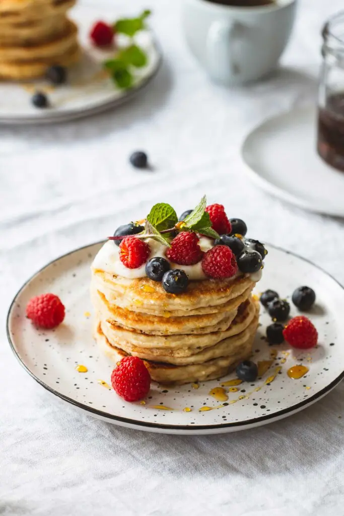 An angled image of a breakfast table setting with a stack of whole wheat vegan pancakes topped with vegan yoghurt and berries in the foreground. Another stack sits in the background next to a cup of coffee and a pot of maple syrup. 
