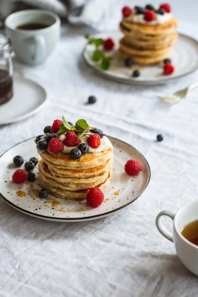 Image of a breakfast table setting on a linen white tablecloth. A stack of whole wheat vegan pancakes sits on a white plate in the front topped with yoghurt and fresh berries. Another plate site in the background with coffee cups and maple syrup. 