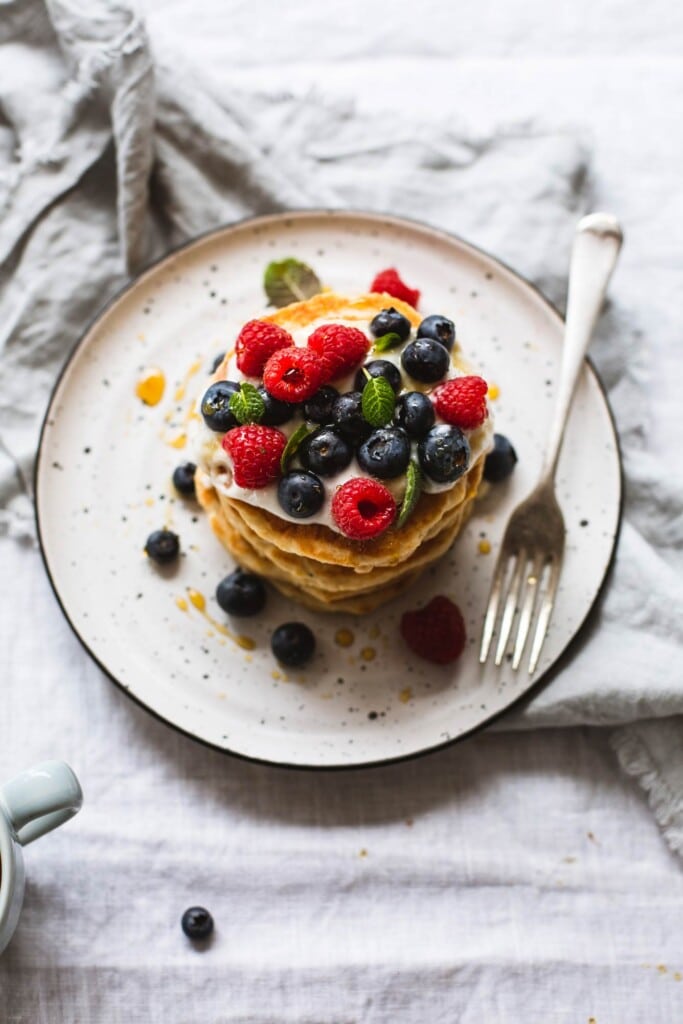 An over-head image of a stack of whole wheat vegan pancakes topped with vegan yoghurt and fresh berries sitting on a white plate on a crinkled linen tablecloth. 