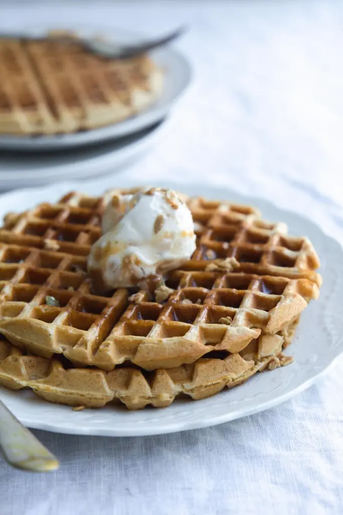 A close-up image of a serving of vegan pumpkin waffles on a white plate with ice-cream, granola sprinkles and maple syrup