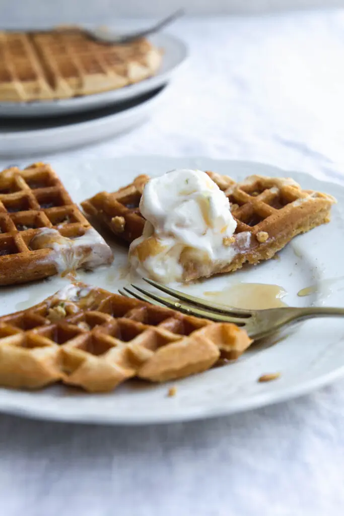 Close-up image of a vegan pumpkin waffles with melting ice-cream and maple syrup partially eaten on a white plate. 