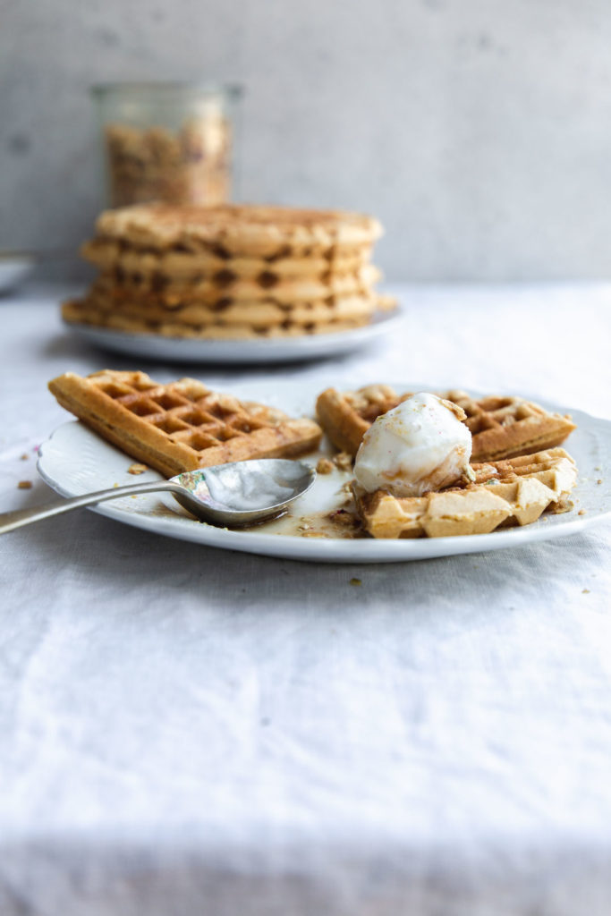 A table top image from the front of a plate holding a partially eaten serve of vegan pumpkin waffles with ice-cream and maple syrup. A stack of pancakes and a jar of granola sit in the background on a crinkled linen tablecloth. 