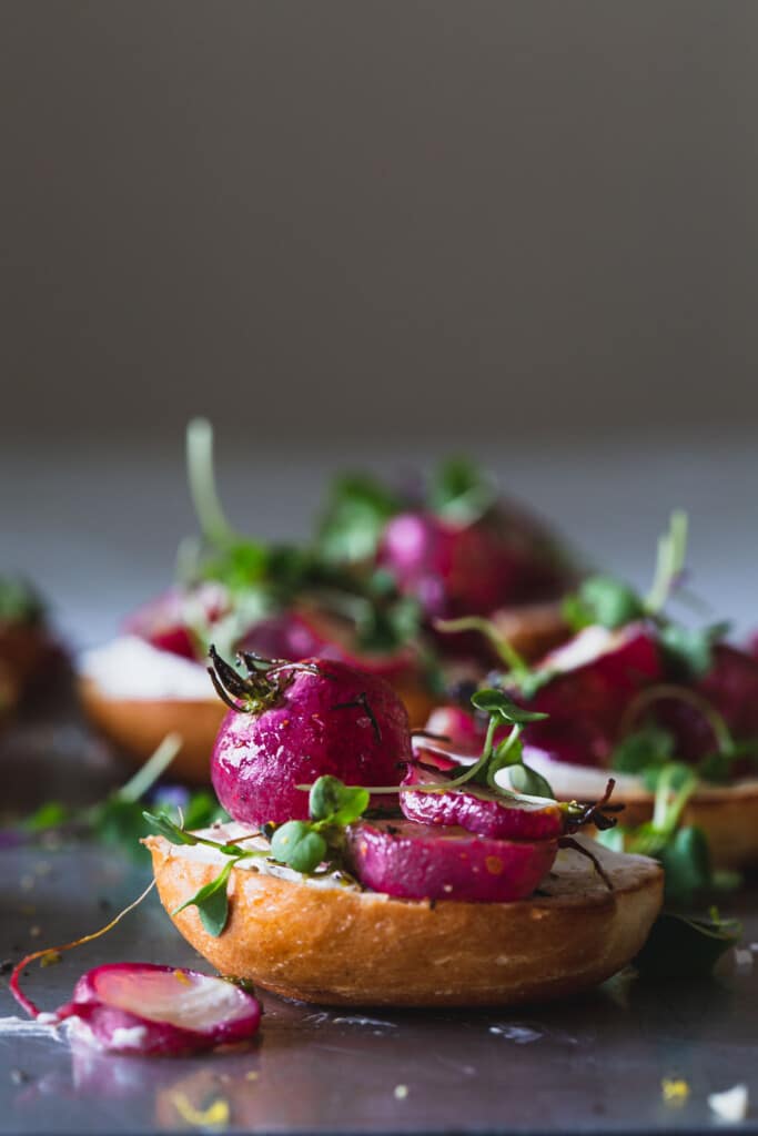 A front on image of a tray of roasted radishes sitting on mini bagels with cream cheese and micros greens. Front bagel in focus. 