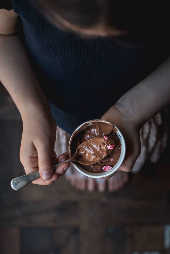An overhead image of a small girl eating a pot of vegan chocolate mousse. The shot is taken above her head so we can see all the way down to her bare feet. 