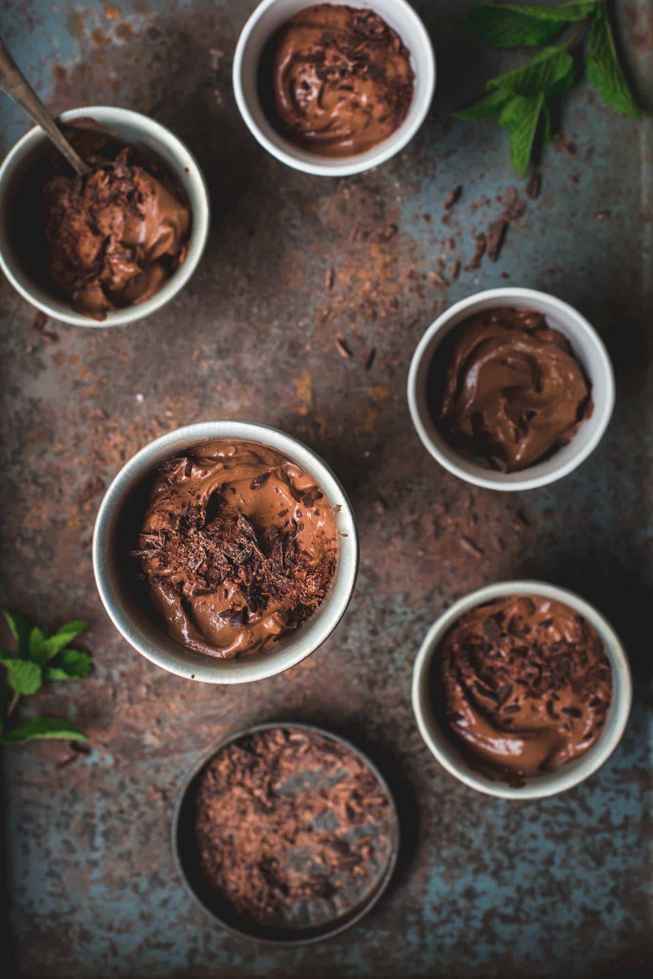 5 white ramekins full of vegan mousse sit on a vintage baking try with mint leaves scattered around. One ramekin is slightly elevated towards to the camera and is the focus point
