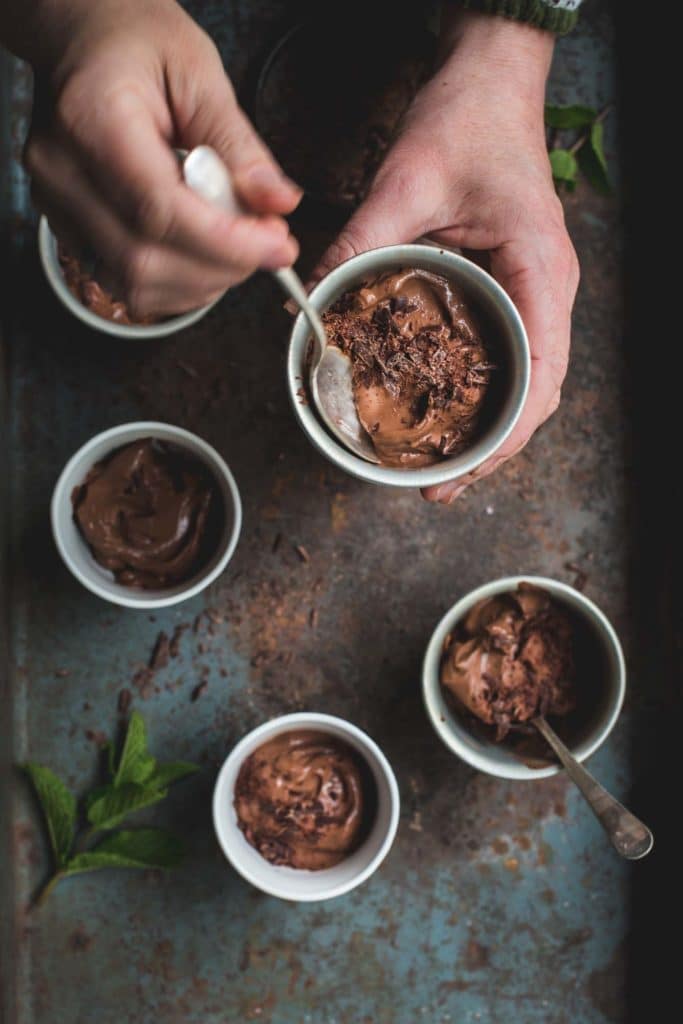 An overhead image of 5 pots pots of vegan mousse sitting on a vintage baking tray with one pot being held by a woman's hands. She is scooping a spoon of the mousse with a spoon. 