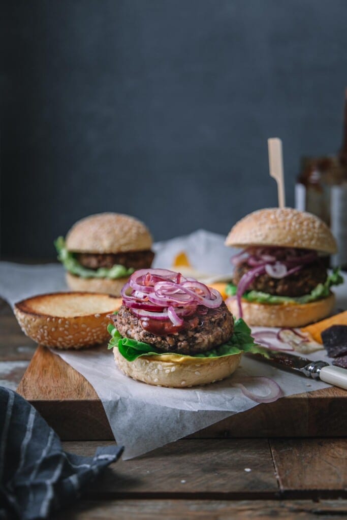 Image shot from the front of 3 BBQ vegan black bean burgers on a wooden board with parchment payment on top. The front centre burger is in focus and has its top burger bun set aside to show the burger, BBQ sauce and fried onions on top.