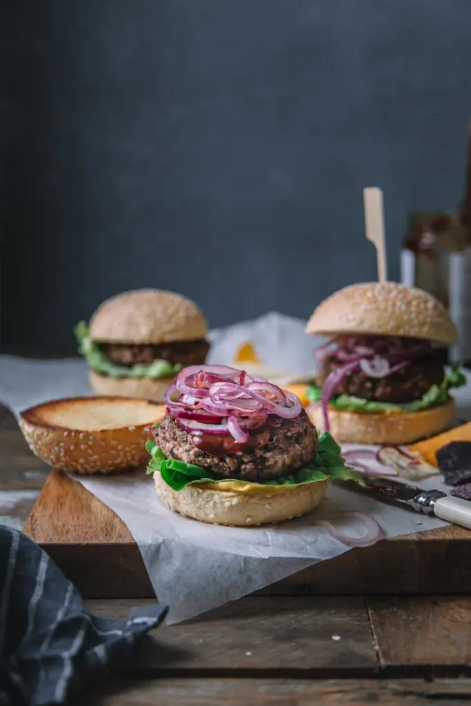 Image shot from the front of 3 BBQ vegan black bean burgers on a wooden board with parchment payment on top. The front centre burger is in focus and has its top burger bun set aside to show the burger, BBQ sauce and fried onions on top. 