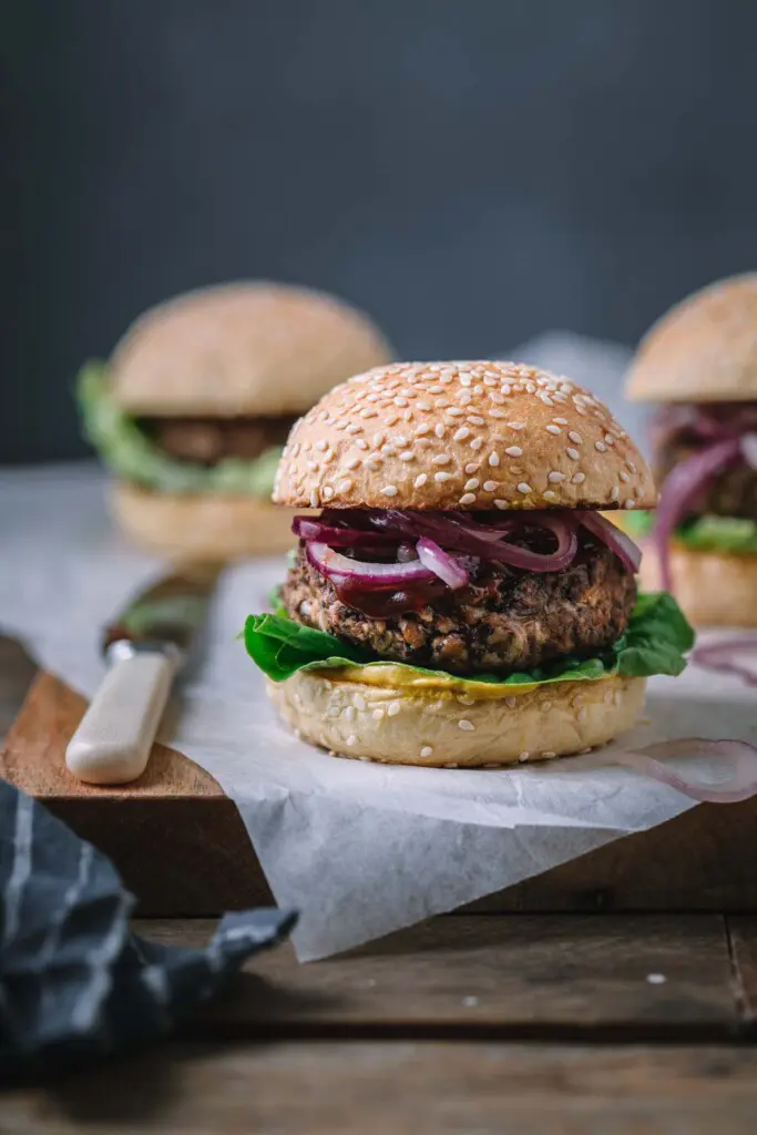 Front on image of a BBQ Vegan Black Bean Burger sitting on a rustic wooden board with two burgers in the background