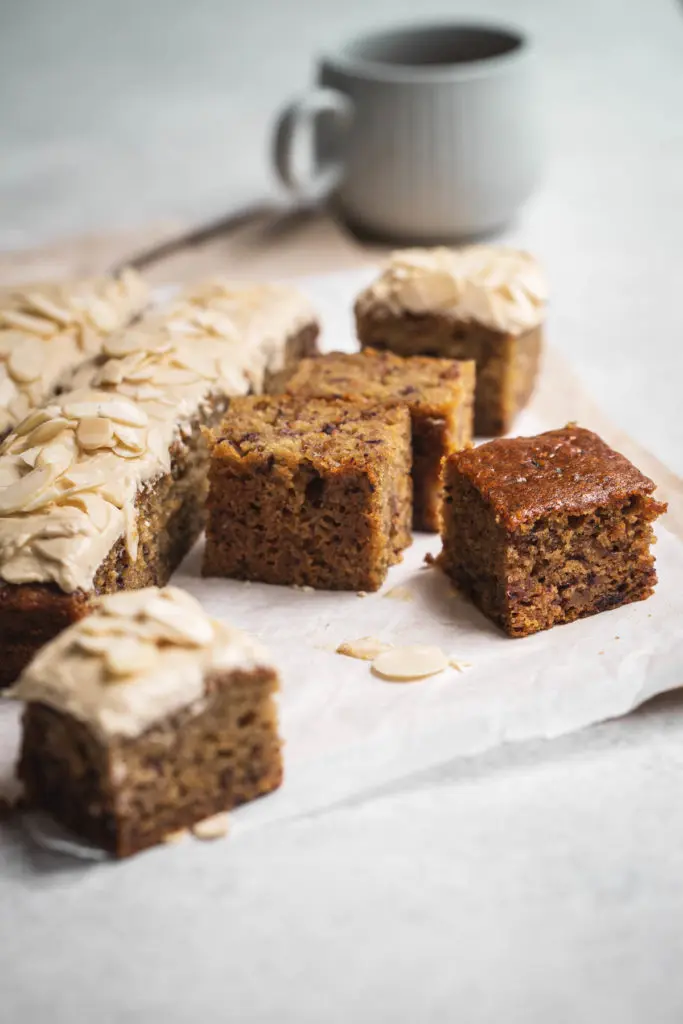 A frosted sticky date cake partially cut in to squares sitting on textured parchment paper with a cup in the background. 