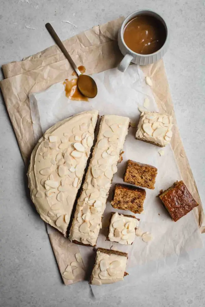 An overhead shot of a frosted round sticky date cake partially cut in to squares sitting on textured baking paper. Only half the cake is cut and some of the squares are sitting sideways to show the texture of the finished cake. A cup of salted caramel with a spoon sits to the top of the frame. 
