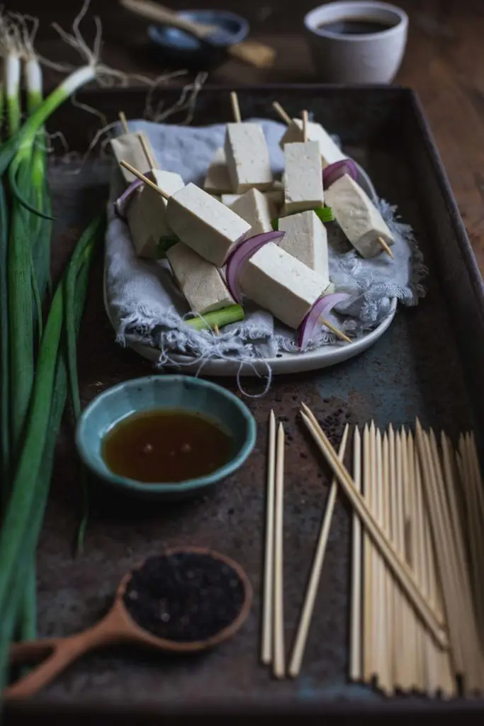 A plate of the pre-cooked tofu yakitori skewers, a pinch bowl of oil, a pot of dipping sauce and other ingredients sit inside a rustic tray ready to be grilled. 