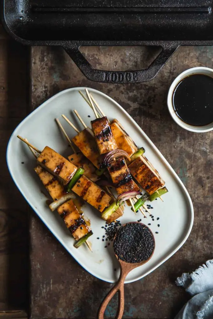 The finished grilled tofu yakitori skewers sit stacked on a rectangle white serving plate with a small bowl of dipping sauce nearby. 