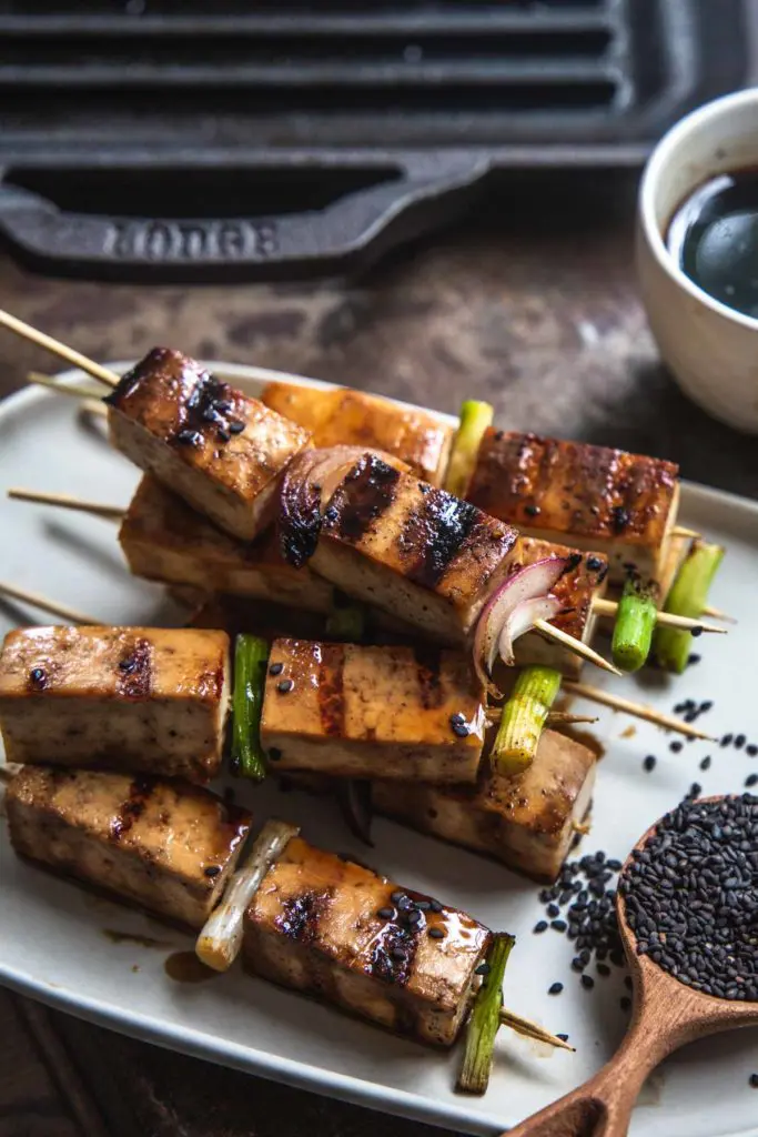 A close up image of a heaped pile of grilled tofu yakitori skewers sitting on a rectangle white serving plate. The cooking glaze shines on each skewer. 