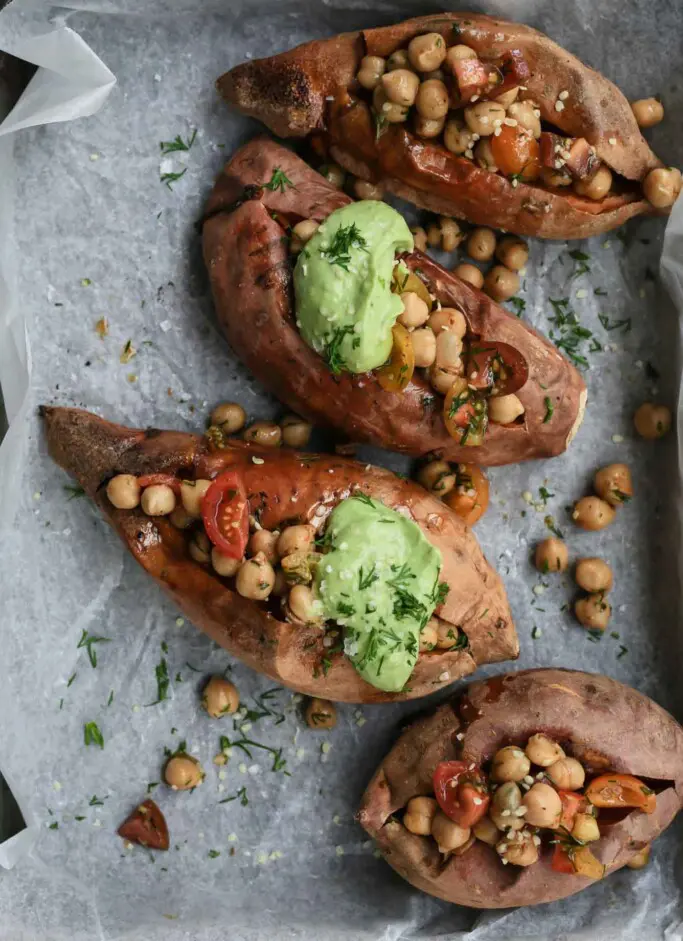 Overhead image of 3 herbed chickpea stuffed sweet potatoes on a baking paper served with a big dollop of green avocado tahini dressing. 