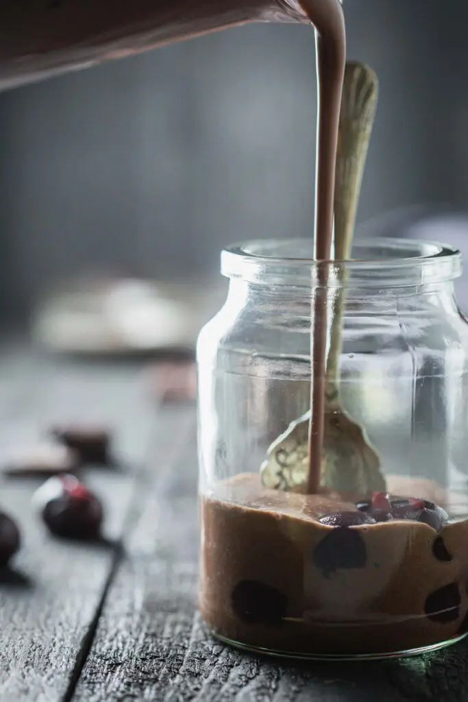 A chocolate drink being poured over cherries in a jar. 