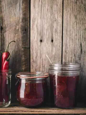 Jars of jam and chillies sitting on a wooden shelf.