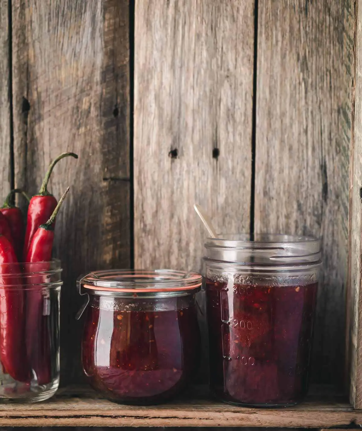 Jars of jam and chillies sitting on a wooden shelf.