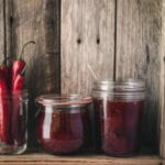 Jars of jam and chillies on a wooden shelf.