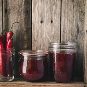 Jars of jam and chillies on a wooden shelf.